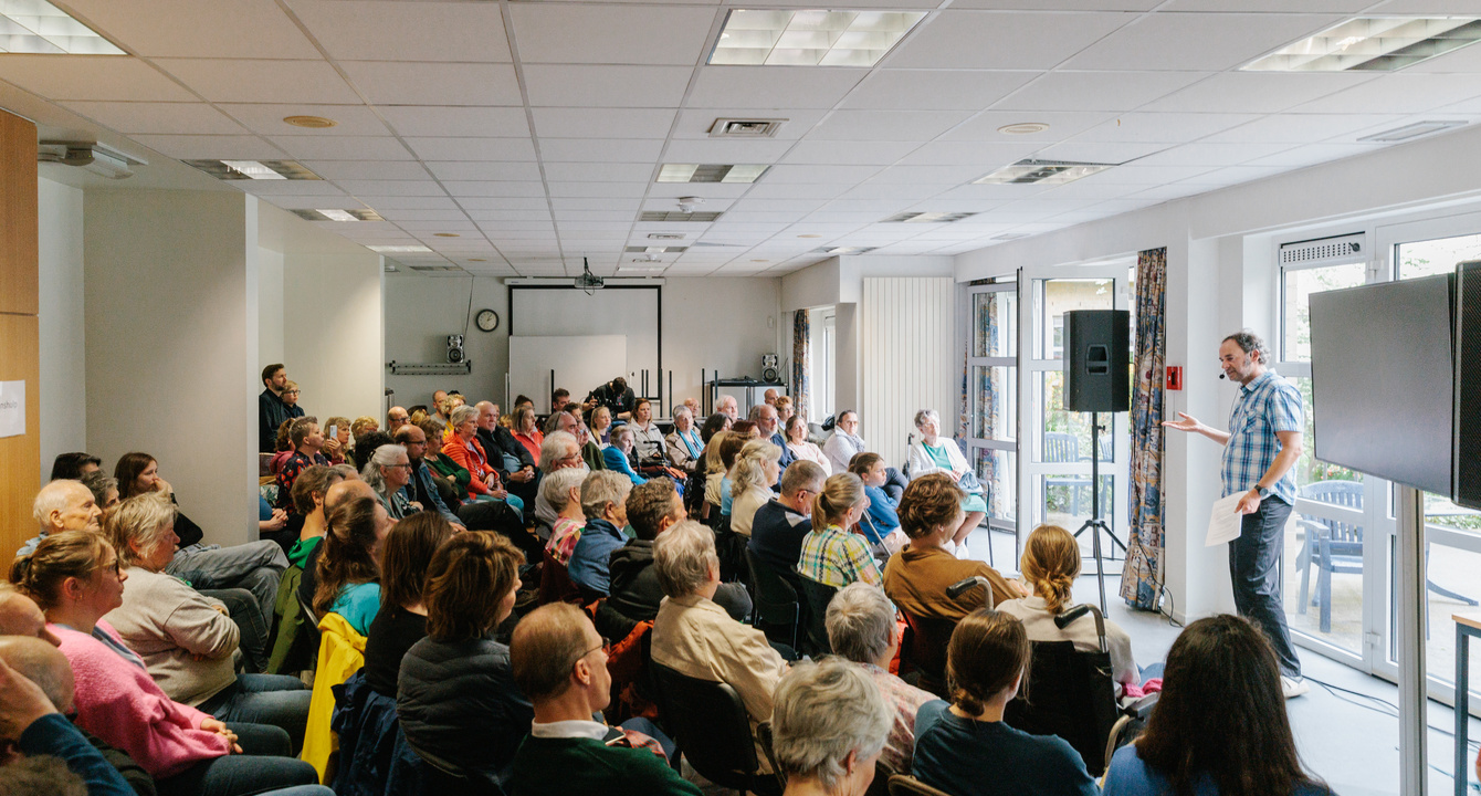 mensen zitten in een zaal, man staat op podium