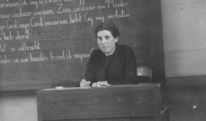 Een vrouw zit achter een lessenaar voor een schoolbord