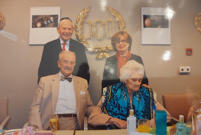 Een oudere man en vrouw zitten aan een tafel. Achter staan een man en een vrouw recht. Op de muur hangt het cijfer 100