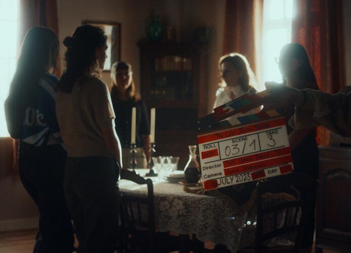 Mensen staan rond tafel. Filmbord vooraan.