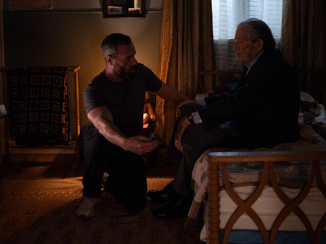 Man zit gehurkt naast bed waarop andere man zit